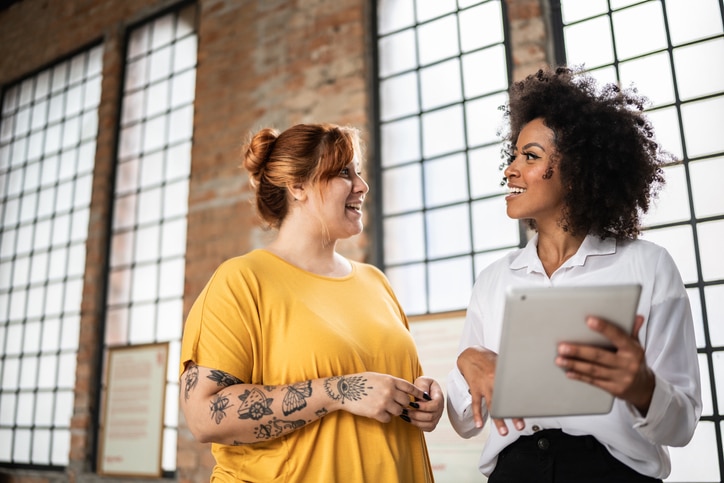 Two female coworkers talking. One is a white woman with tattoos, and one is a Black woman with an afro, holding a tablet.